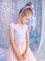 A young girl with blonde hair in a bun and a large lavender ribbon bow, wearing a light blue and pink Barbie printed leotard with a pink tulle skirt, posing in front of a snowy winter-themed backdrop.