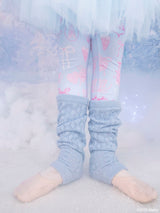 A close-up of a girls legs wearing light blue knit leg warmers over blue Tutu du Monde x Barbie leggings with pink ballet-themed Barbiep rints, standing in a snowy, dreamy studio setting.
