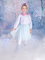 A young girl modeling a light blue Barbie in The Nutcracker skirt and jumper with pink accents, standing in a blue, icy forest setting with glowing snowflakes and mist.