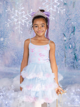 A young girl wearing a light blue and pink Barbie x Tutu du Monde tutu dress with a matching headband and tiara, standing in front of a snowy, icy backdrop with lights.
