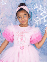 A young girl wearing a pink Barbie ballet top with puff sleeves, a matching pink Barbie headband, and a sparkly heart skirt, posing against a blue and white icy background.