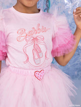 A close-up of a child wearing a pink Barbie-themed tutu dress with a tulle top, pink ruffled sleeves, a bow-tie ballet shoe graphic, and a heart-shaped embellishment at the waist.